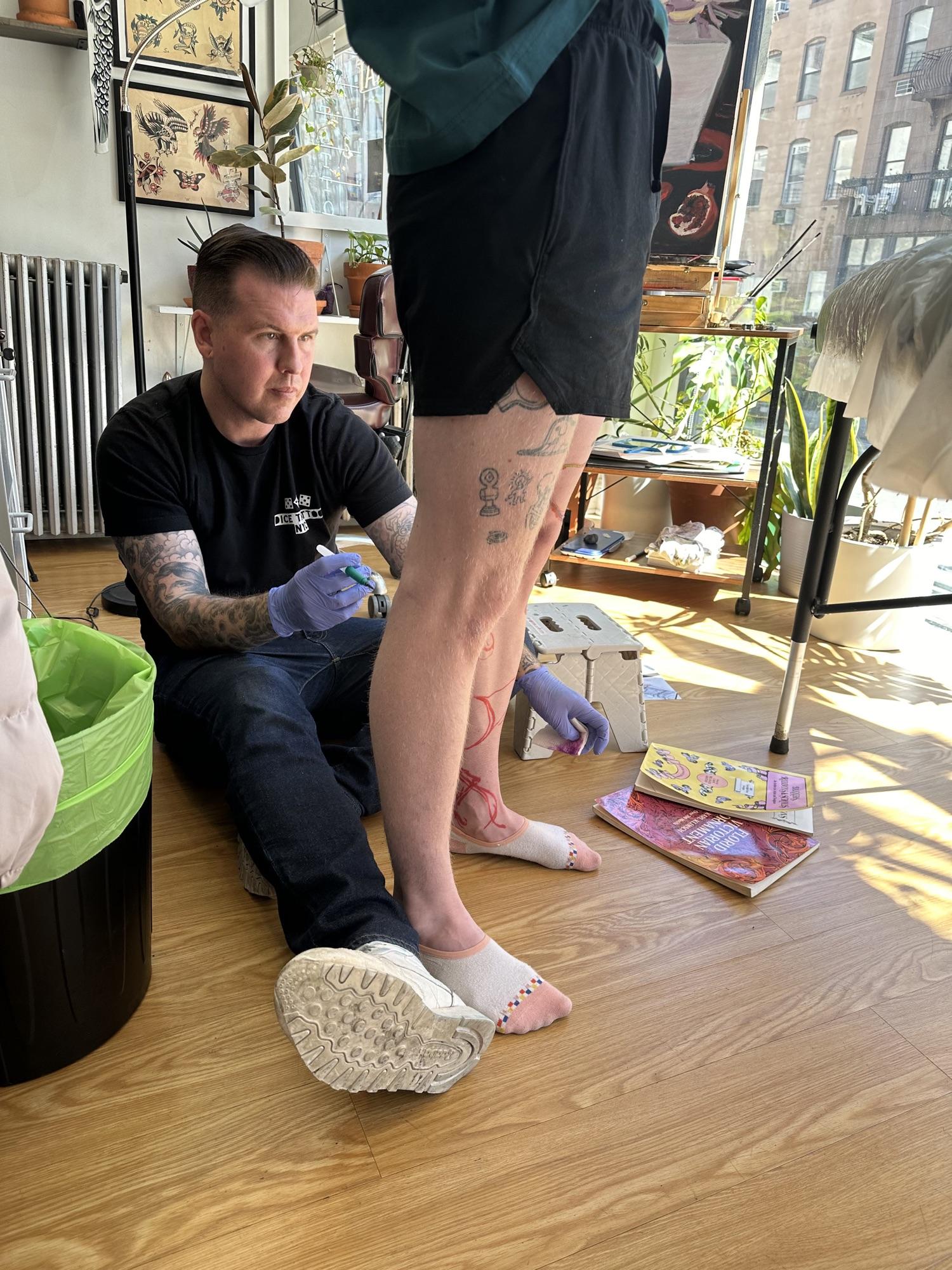 Mike Stapleton sitting on the studio floor, drawing a design onto a client’s leg